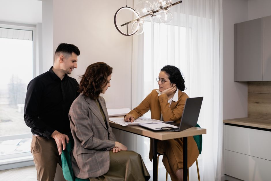 Professional real estate agent gesturing towards a laptop screen while speaking with a homeowner in a bright, modern livin...